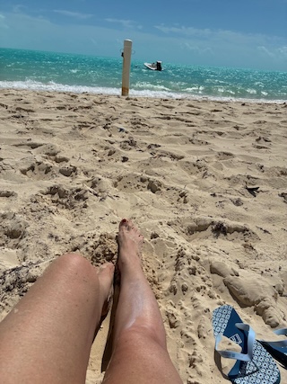 The author's partner's sun-bronzed legs lay on Long Bay beach pointing towards a wooden mooring post on the sea's edge